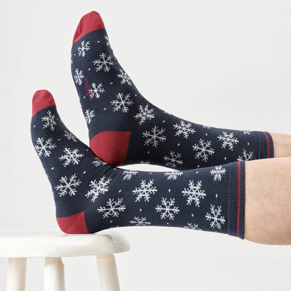 Navy socks with white snowflake pattern and red accents worn by a person sitting on a white stool.