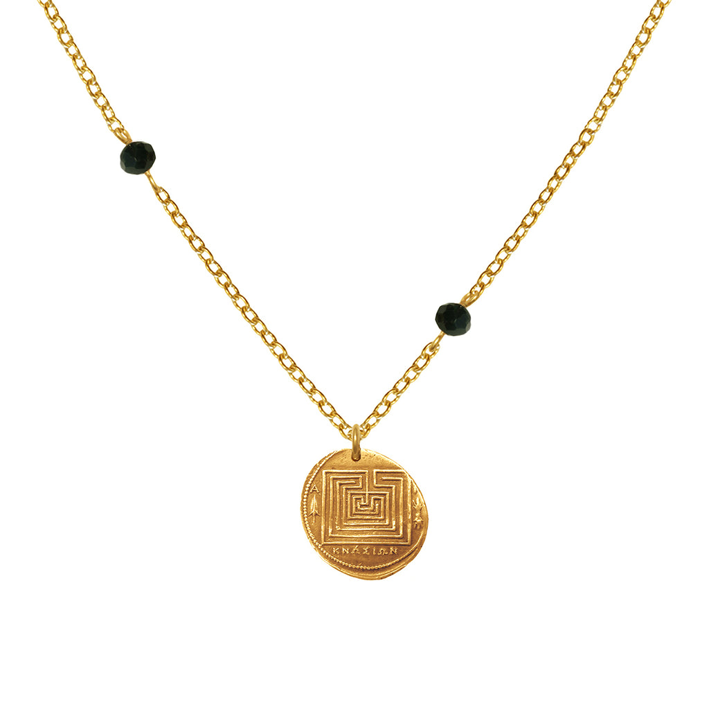A gold necklace featuring a coin-shaped pendant with a geometric pattern, suspended from a chain link, with two small black beads on either side of the pendant.