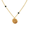 A gold necklace featuring a coin-shaped pendant with a geometric pattern, suspended from a chain link, with two small black beads on either side of the pendant.